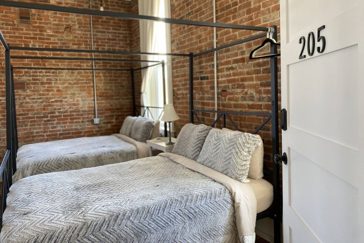Una habitación con dos camas, una pared de ladrillo, una puerta con el número 205 y una lámpara colgante.