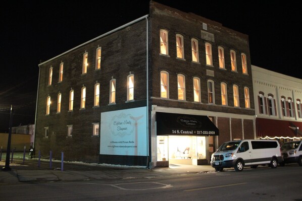 A brick building with illuminated windows and a storefront. - Eighteen-Ninety Sleepover (Casey)