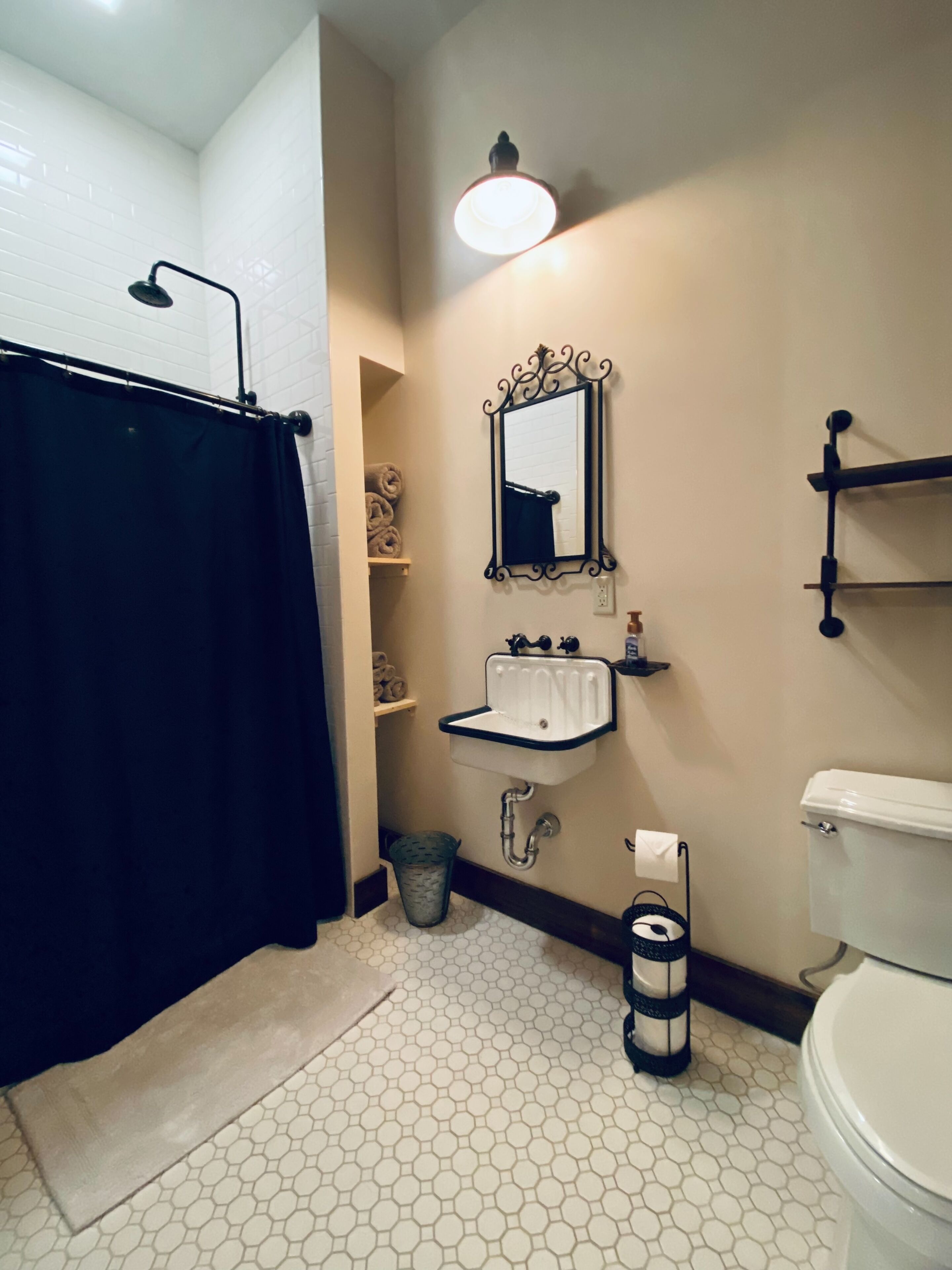 Un baño con cortina de ducha negra, lavamanos blanco con espejo arriba, inodoro y cubo de basura.