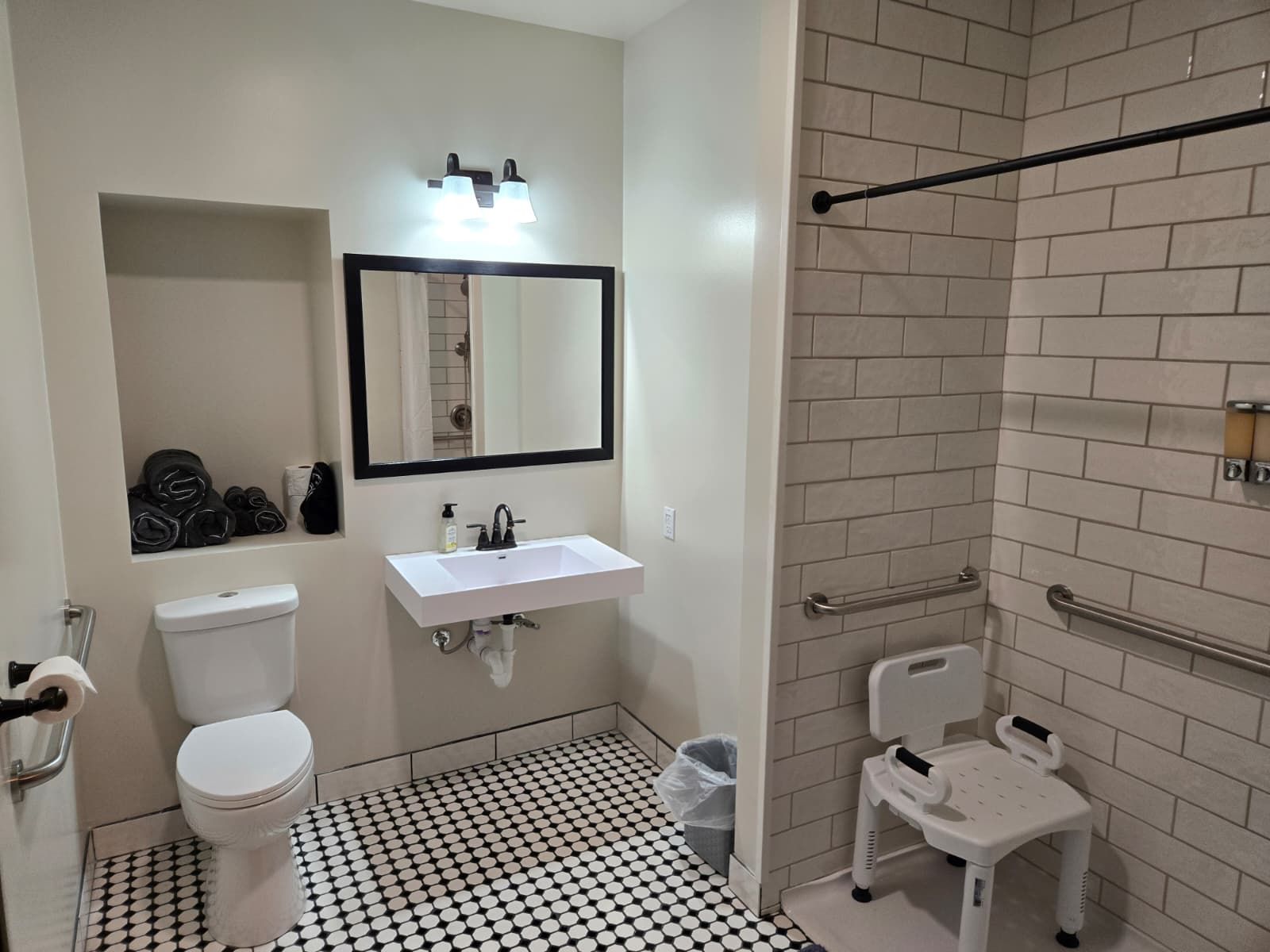 A bathroom with a white toilet, a sink with a mirror above it, a shower area with a handrail, and a small white chair.