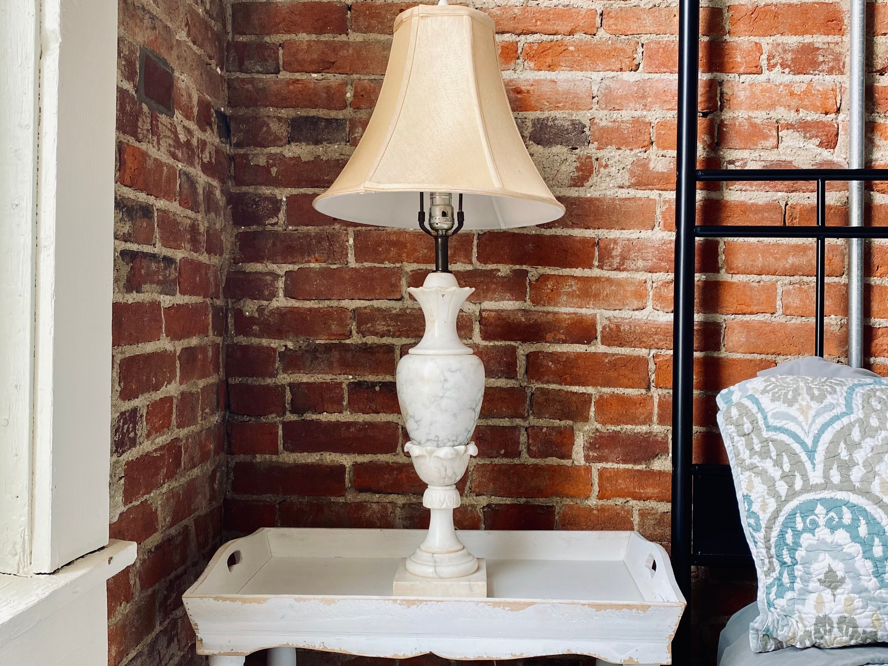 Una lámpara de mesa blanca con pantalla color beige sobre una mesita blanca, frente a un muro de ladrillo.