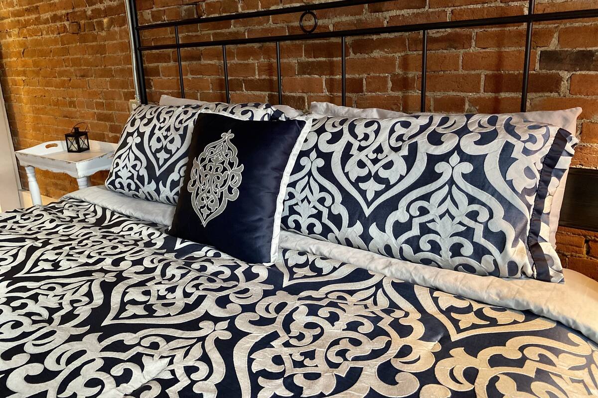 Una cama con una colcha y almohadas estampadas, una mesita de noche blanca y una pared de ladrillo al fondo.