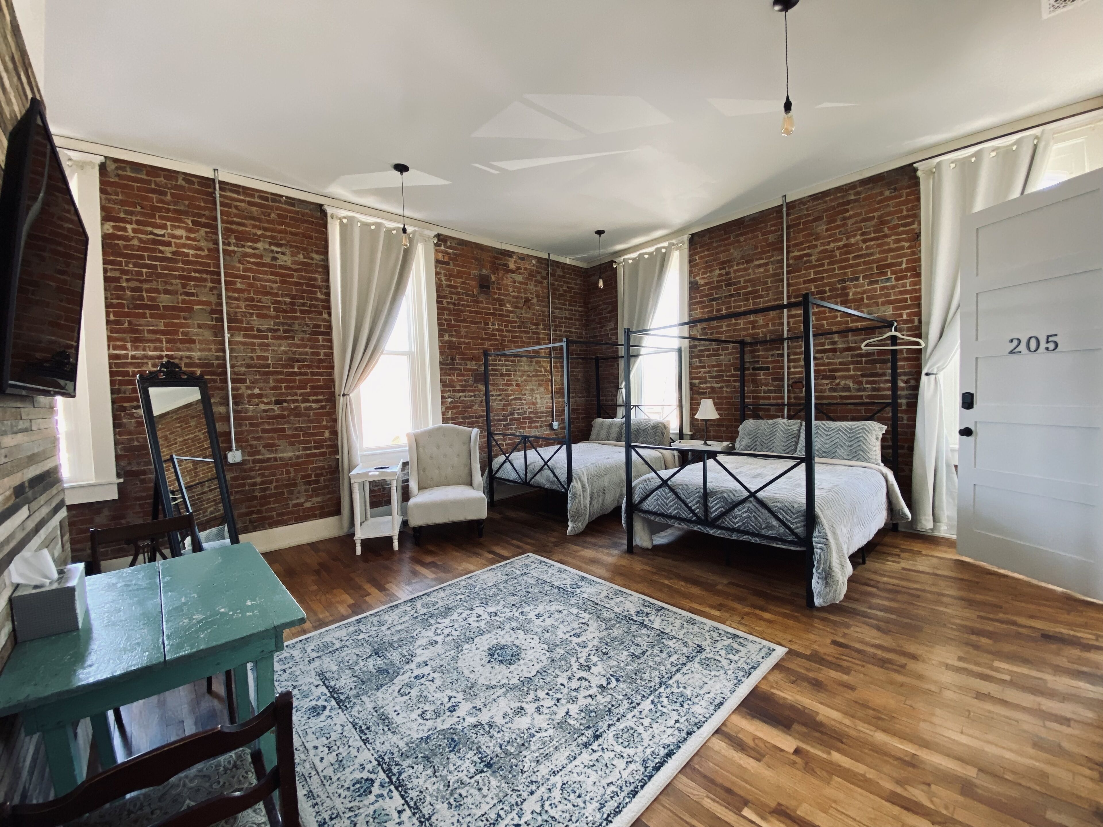 Una habitación con pared de ladrillo, una cama con dosel, un escritorio con silla, una lámpara colgante y una puerta con el número 205.