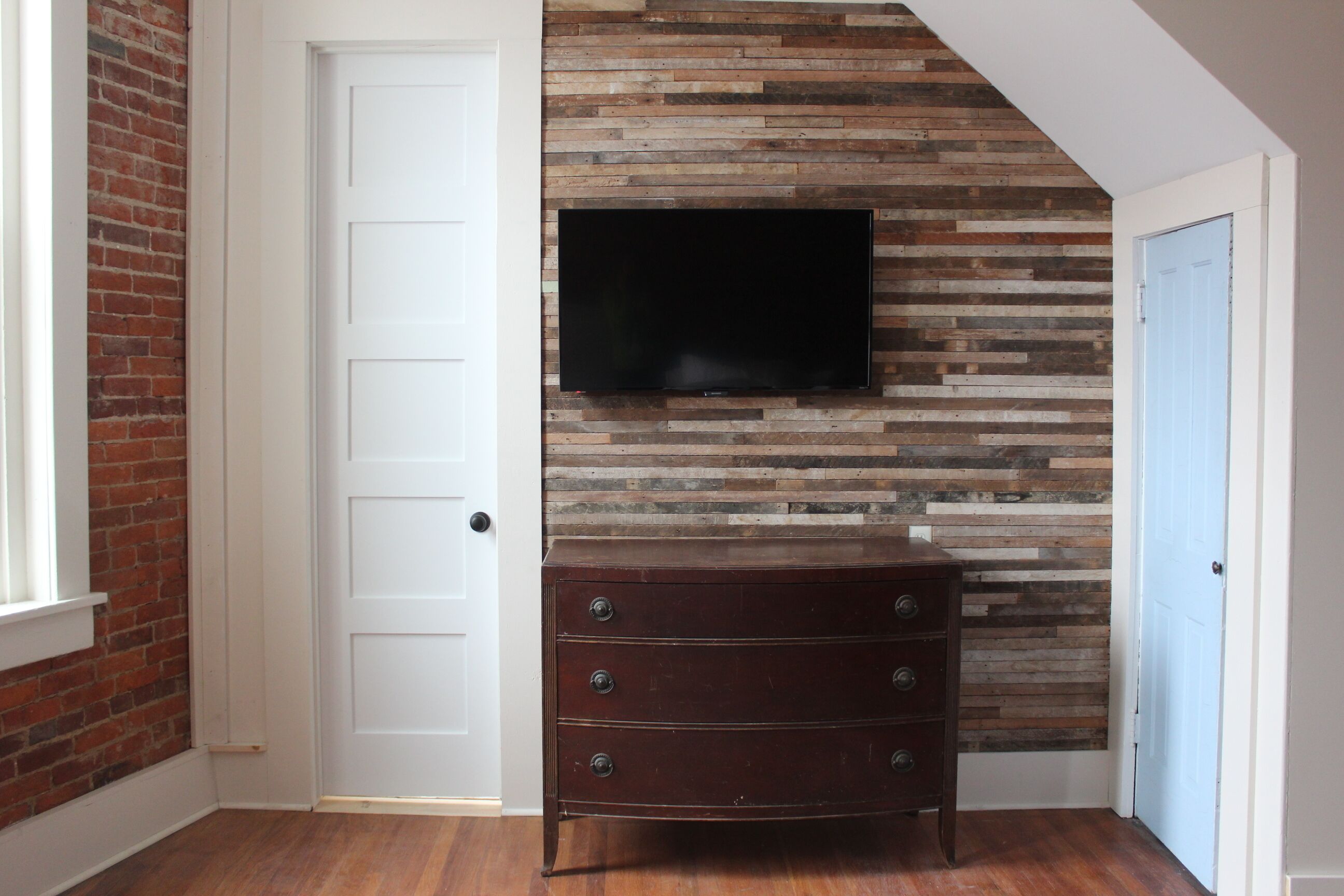 A room with a brick wall, a wooden dresser, and a flat-screen TV mounted on the wall.