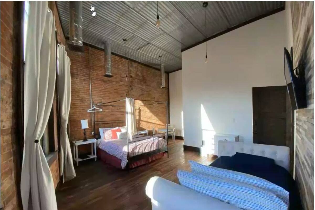 Un dormitorio moderno con una cama grande, una cama más pequeña y un sofá. Se observa una pared de ladrillo y un techo blanco.