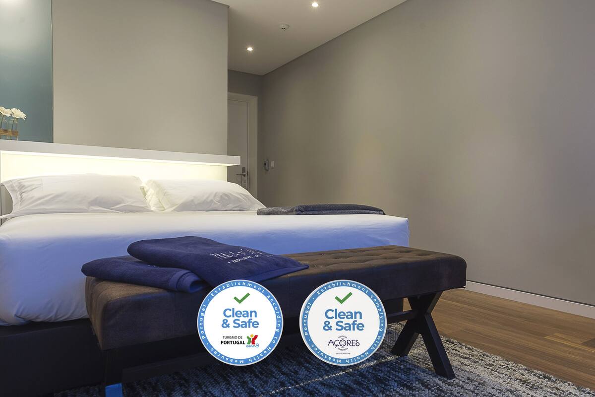 Quarto de hotel moderno com cama bem arrumada, um banco com etiquetas 'Clean & Safe' e vista para a cidade.
