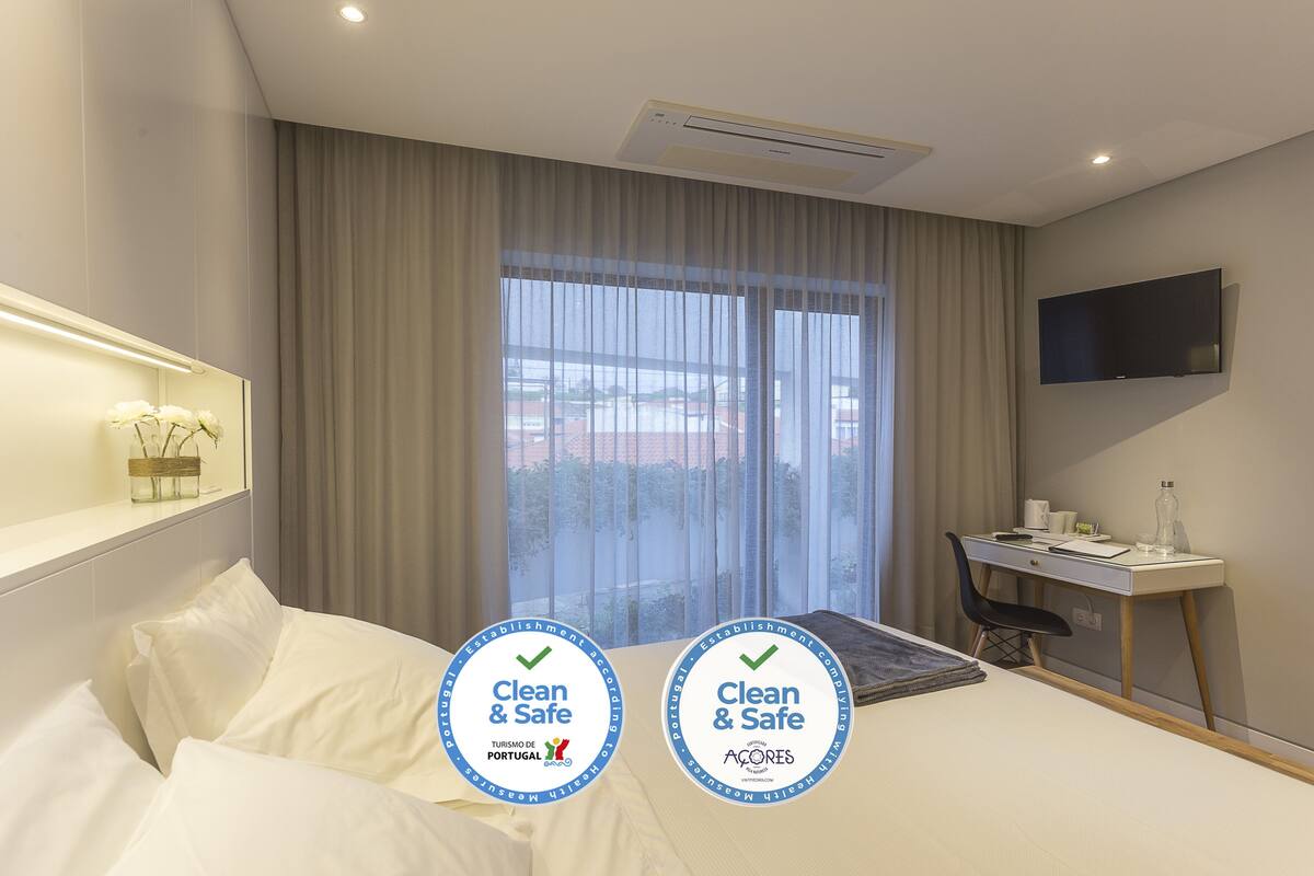 Quarto de hotel com cama, escrivaninha, cadeira, televisão e janela com cortinas.