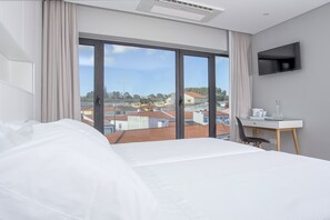 A hotel room with a large bed, a desk with a chair, a flat-screen TV, and a view of the city through large windows. - Praia de Santos - Exclusive Guest House (Ponta Delgada)