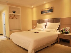 A hotel room with a large bed, a bedside table with a lamp, a nightstand, a wall-mounted shelf with a plant, and a painting above the bed.