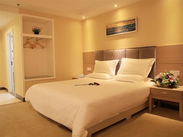 Quarto de hotel com uma cama grande, mesa de cabeceira com luminária, criadeira, prateleira na parede com uma planta e uma pintura sobre a cama.