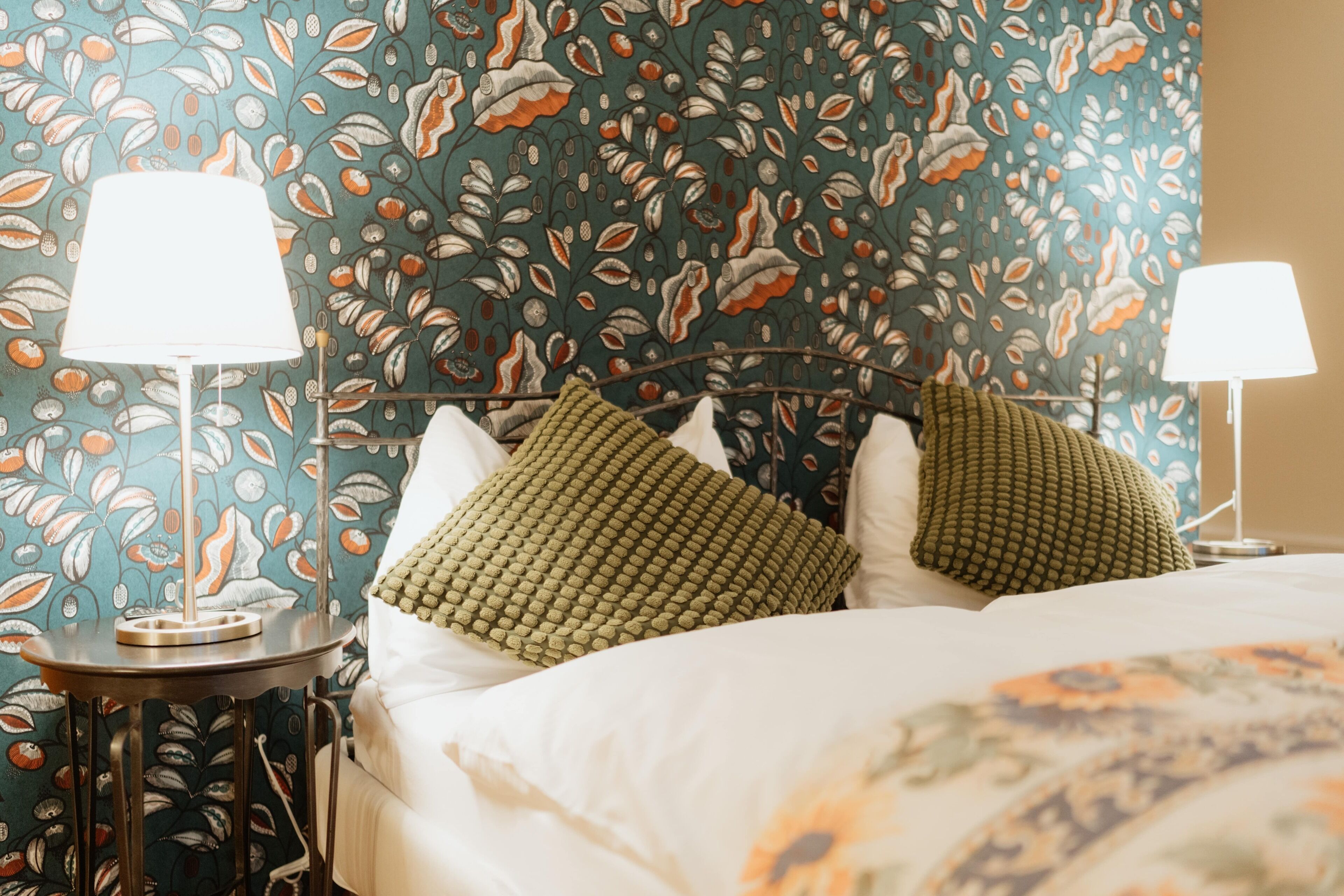 Ein ordentlich bezogenes Bett mit zwei grün gemusterten Kissen, einer weißen Decke und einem blumengemusterten Kopfteil. An jeder Seite des Bettes stehen je eine Nachttischlampe.