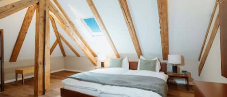 A bedroom with a sloped ceiling, wooden beams, a large bed, bedside tables, a bench, and a small window.