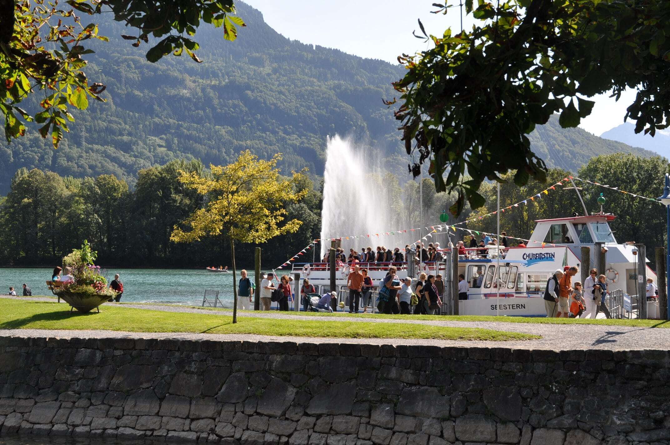 Un bateau était amarré à un quai au bord du lac, des personnes s’étaient rassemblées autour, une fontaine se trouvait à l’arrière-plan et un paysage montagneux s’étendait à perte de vue.