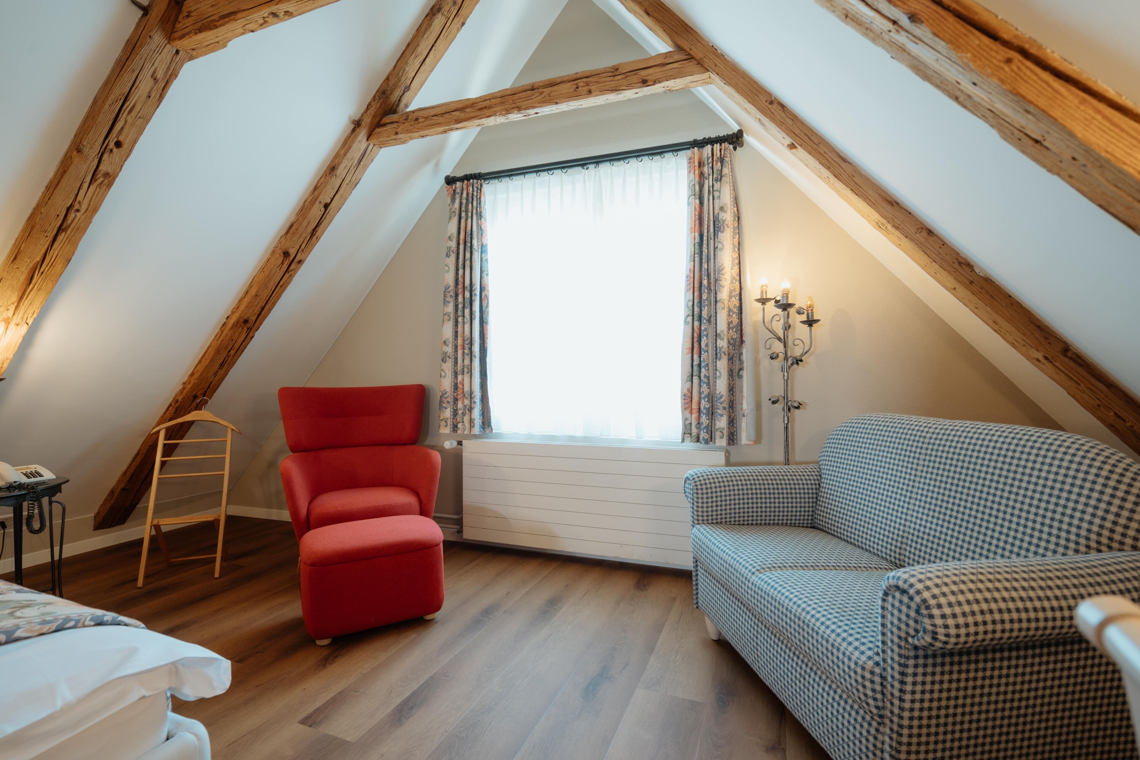 Una camera da letto mansardata accogliente, con soffitto spiovente, una sedia rossa, un divano a quadri e una finestra con tende.