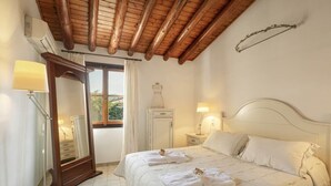 A bedroom with a wooden ceiling, a large bed with white linens, a bedside table with a lamp, a wardrobe, and a mirror. - Olympia Village (Samos)