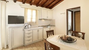 A kitchen with white cabinets, a wooden ceiling, a flat-screen TV, a coffee maker, and a dining table with a pumpkin and squash. - Olympia Village (Samos)
