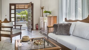 A cozy living room with a wooden sofa, a glass coffee table, and a view of an outdoor patio. - Emily Villas (Kefalonia)