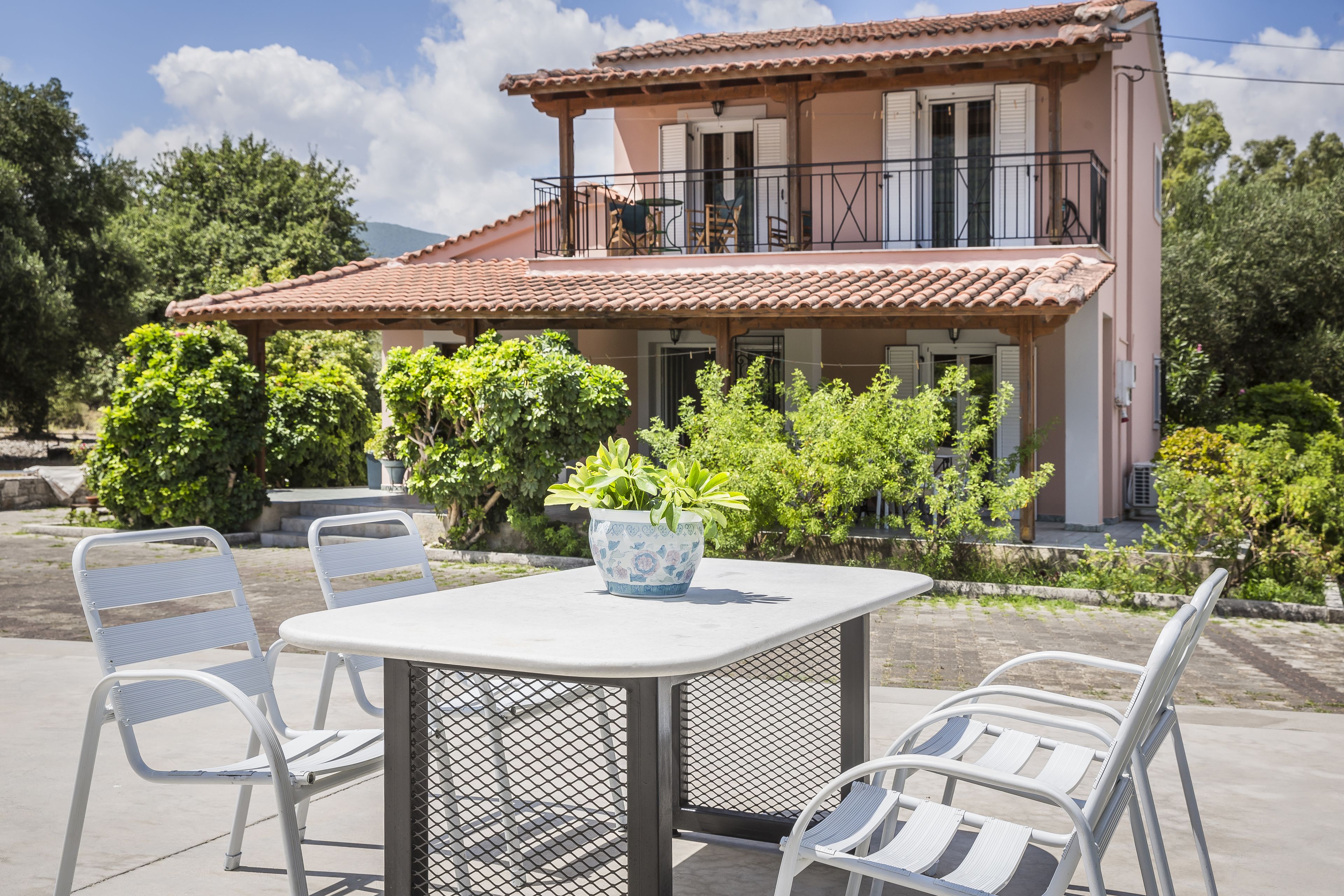 A two-story house with a balcony, a table with chairs, and potted plants.