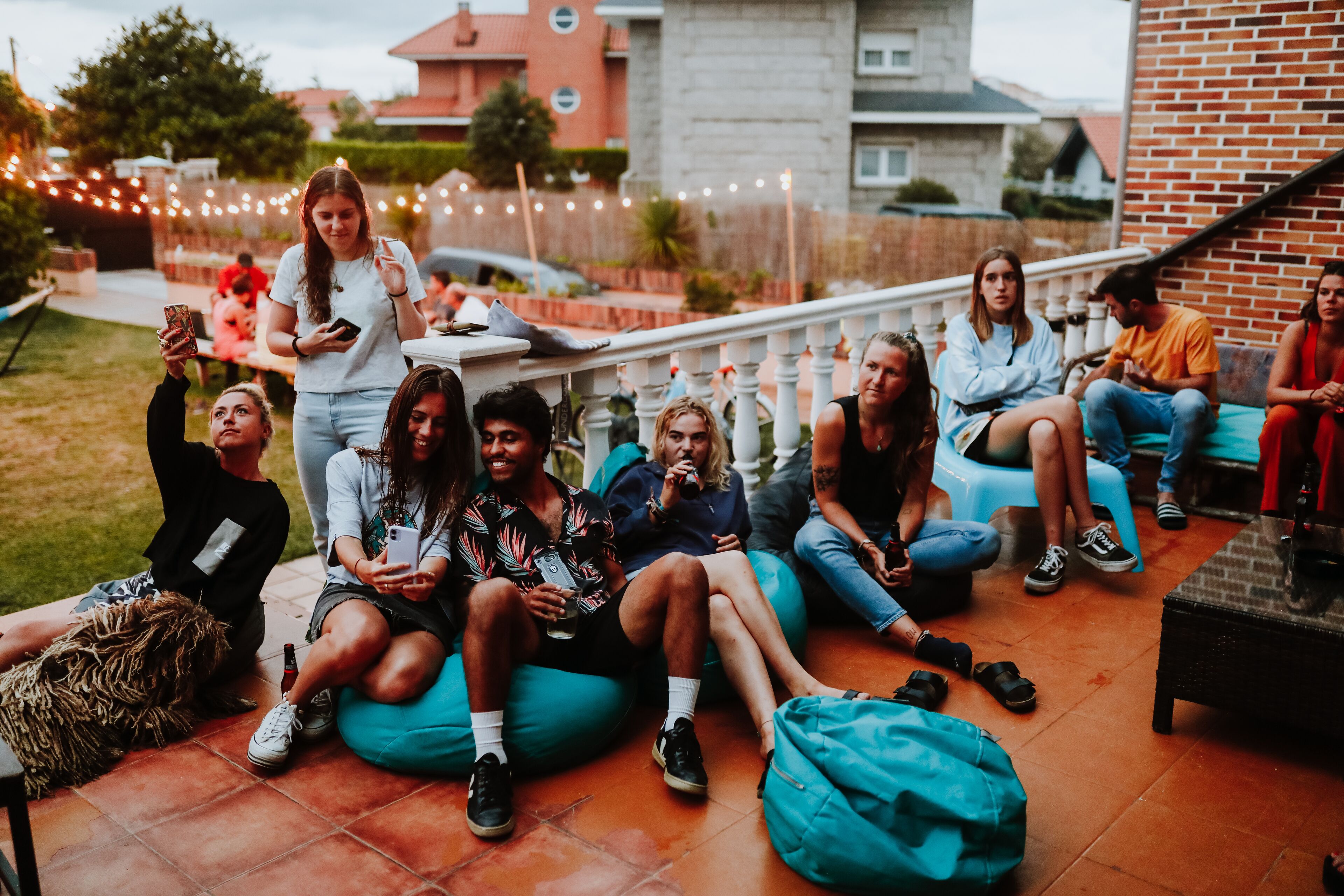 Un grupo de personas sentadas en pufs y sillas en un espacio al aire libre, con una pared de ladrillo y luces colgantes al fondo.