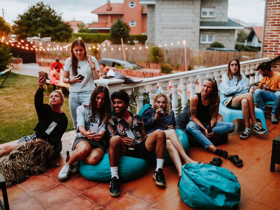 Un grupo de personas sentadas en pufs y sillas en un espacio al aire libre, con una pared de ladrillo y luces colgantes al fondo.