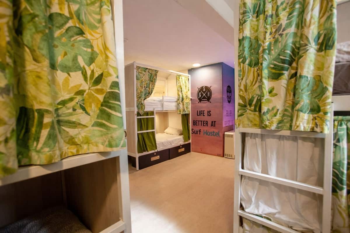 A dormitory room with bunk beds, a green curtain, a wall with a surf-themed poster, and a small air conditioning unit.