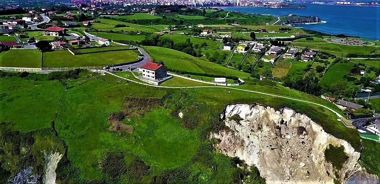 Vista aérea de una zona costera con campos verdes, un acantilado destacado y un edificio de techo rojo.