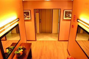 Un área de ascensor con suelo de madera, un banco con flores y cuadros en las paredes.