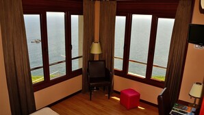 A hotel room with large windows offering a view of the sea, a wooden floor, a bed, a red ottoman, a chair, and a television on the wall. - Hotel La Colina (Gijon)