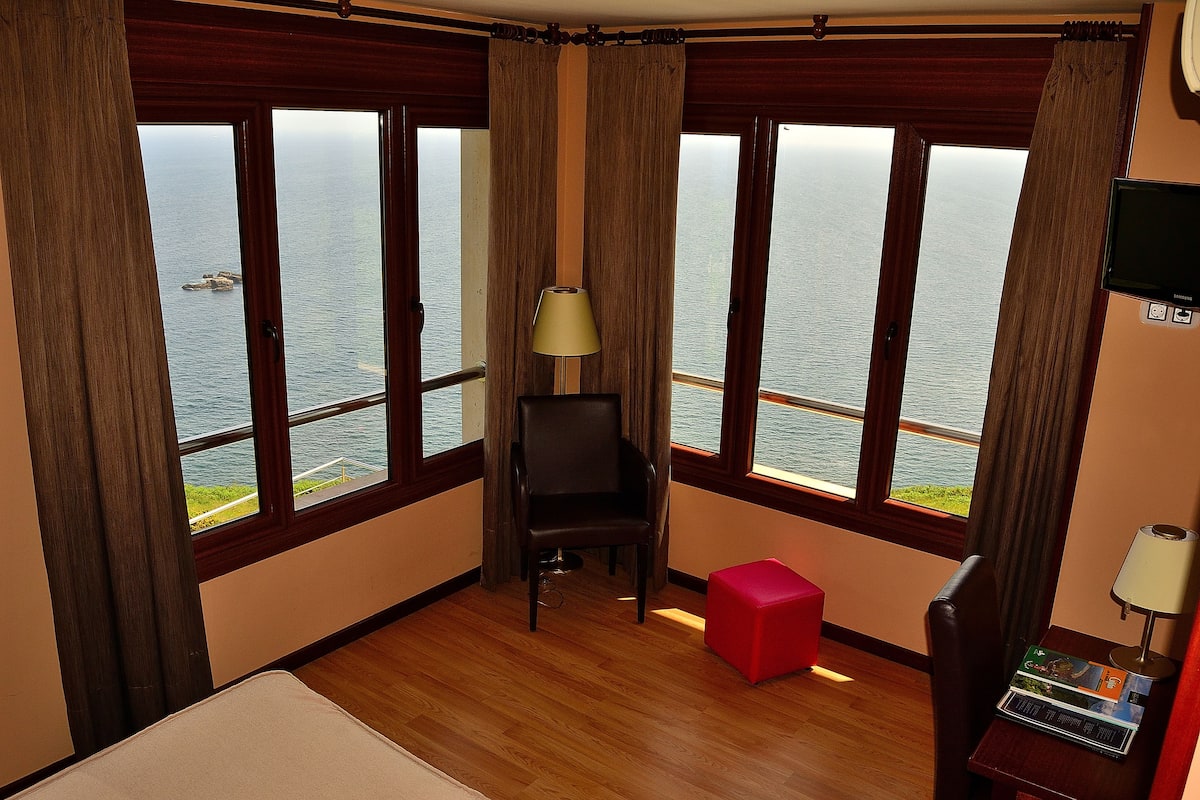 A hotel room with large windows offering a view of the sea, a wooden floor, a bed, a red ottoman, a chair, and a television on the wall.