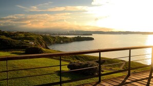 A coastal landscape with a wooden deck overlooking a body of water, grassy hills, and distant city skyline. - Hotel La Colina (Gijon)