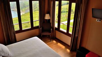 A hotel room with a bed, a chair, a TV, and large windows offering a view of a green landscape.