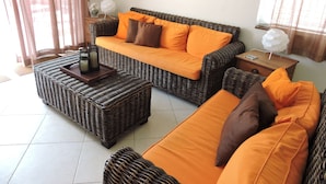 Un salon aménagé avec un ensemble de canapés en osier, des coussins orange, une table d’appoint et une lampe décorative.