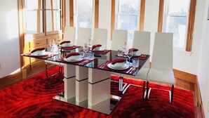 Une salle à manger avec une table en verre dressée pour six personnes, entourée de chaises blanches, agrémentée d’un tapis rouge et de quelques éléments décoratifs en bois.