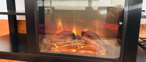 Une cheminée à foyer vitré, avec des bûches et des flammes apparentes, posée sur un plancher en bois.