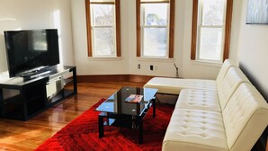 Un salon doté d’un téléviseur à écran plat, d’un canapé blanc, d’un tapis rouge et de trois fenêtres à cadre en bois.