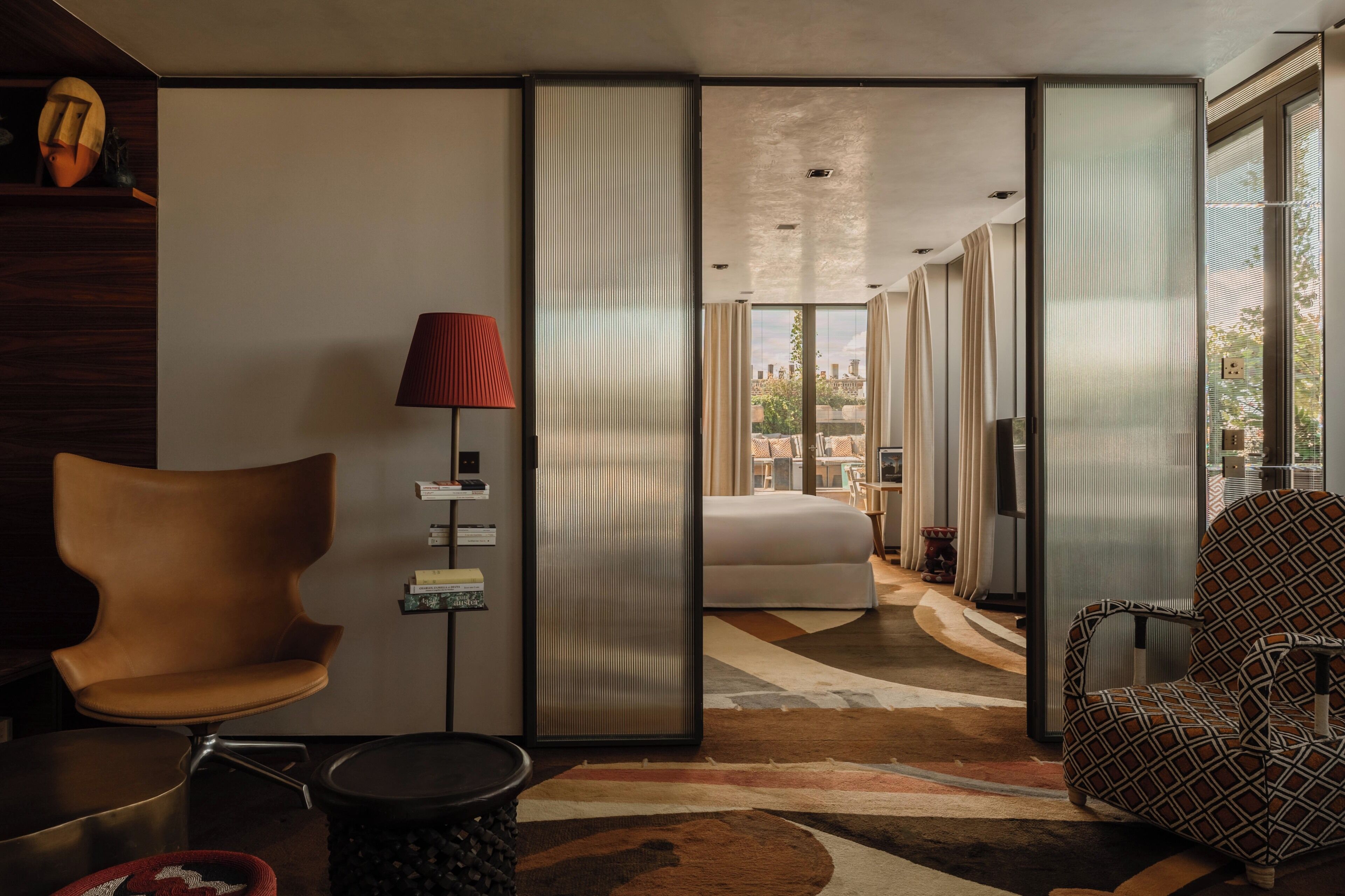 A modern hotel room with a large bed, a sitting area with a chair and a table, a red lamp, and a view of the city through large windows.