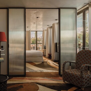 A modern hotel room with a large bed, a sitting area with a chair and a table, a red lamp, and a view of the city through large windows.
