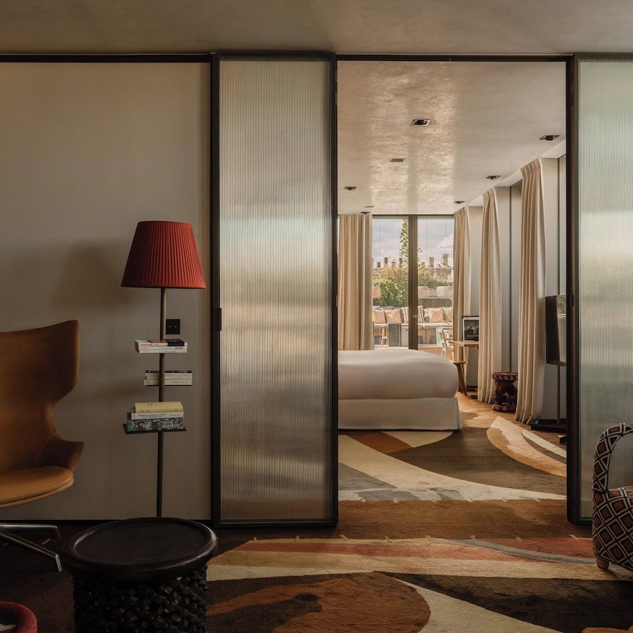 A modern hotel room with a large bed, a sitting area with a chair and a table, a red lamp, and a view of the city through large windows.