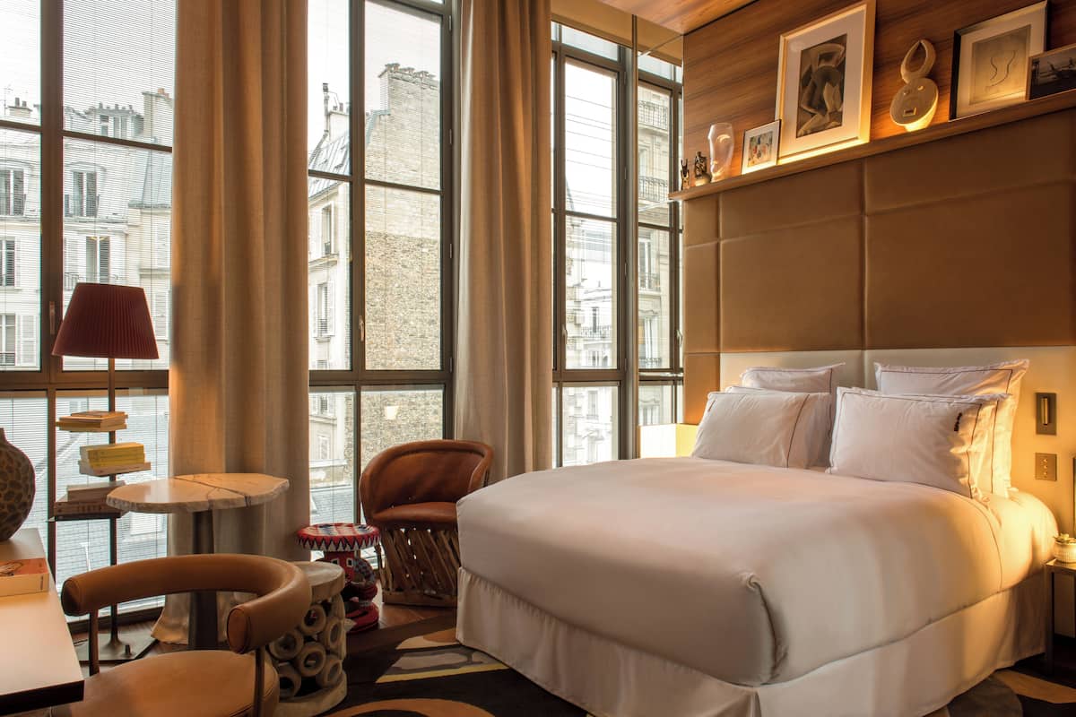 Habitación de hotel con una cama grande, una silla, una mesita y vistas a edificios a través de amplios ventanales.
