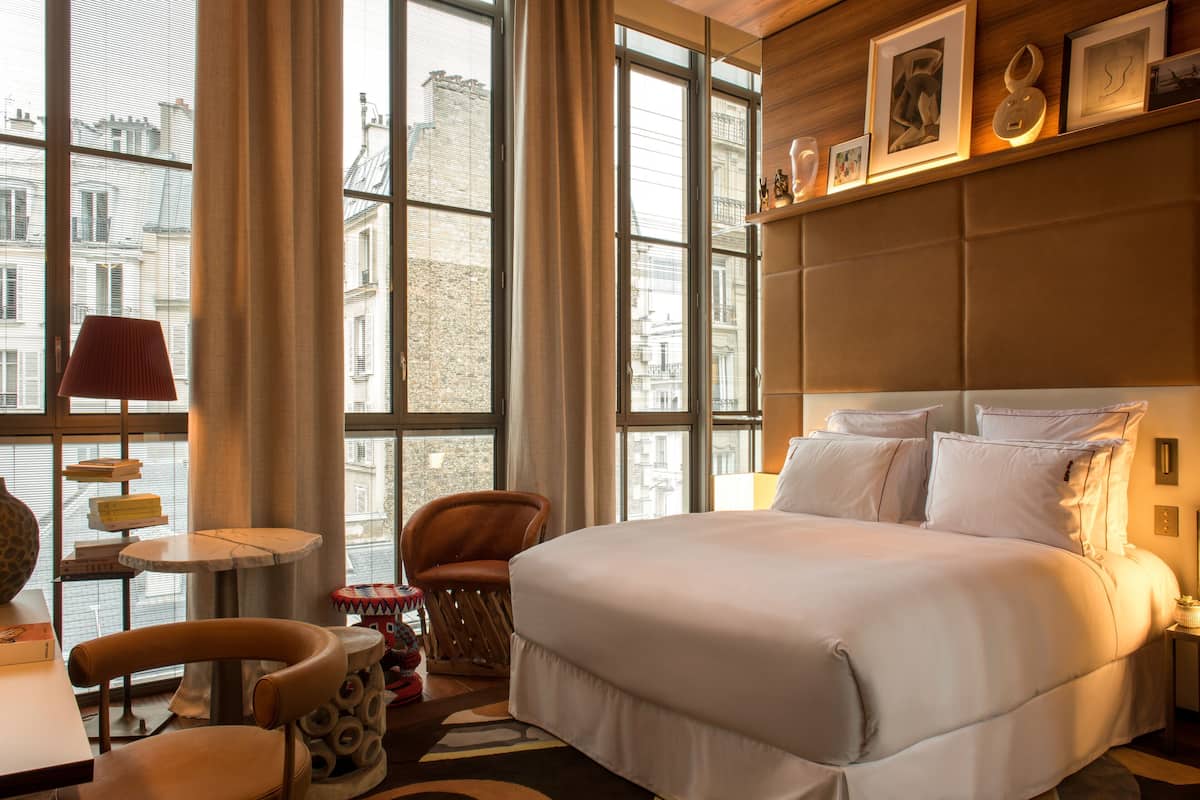 Habitación de hotel con una cama grande, una silla, una mesita y vistas a edificios a través de amplias ventanas.