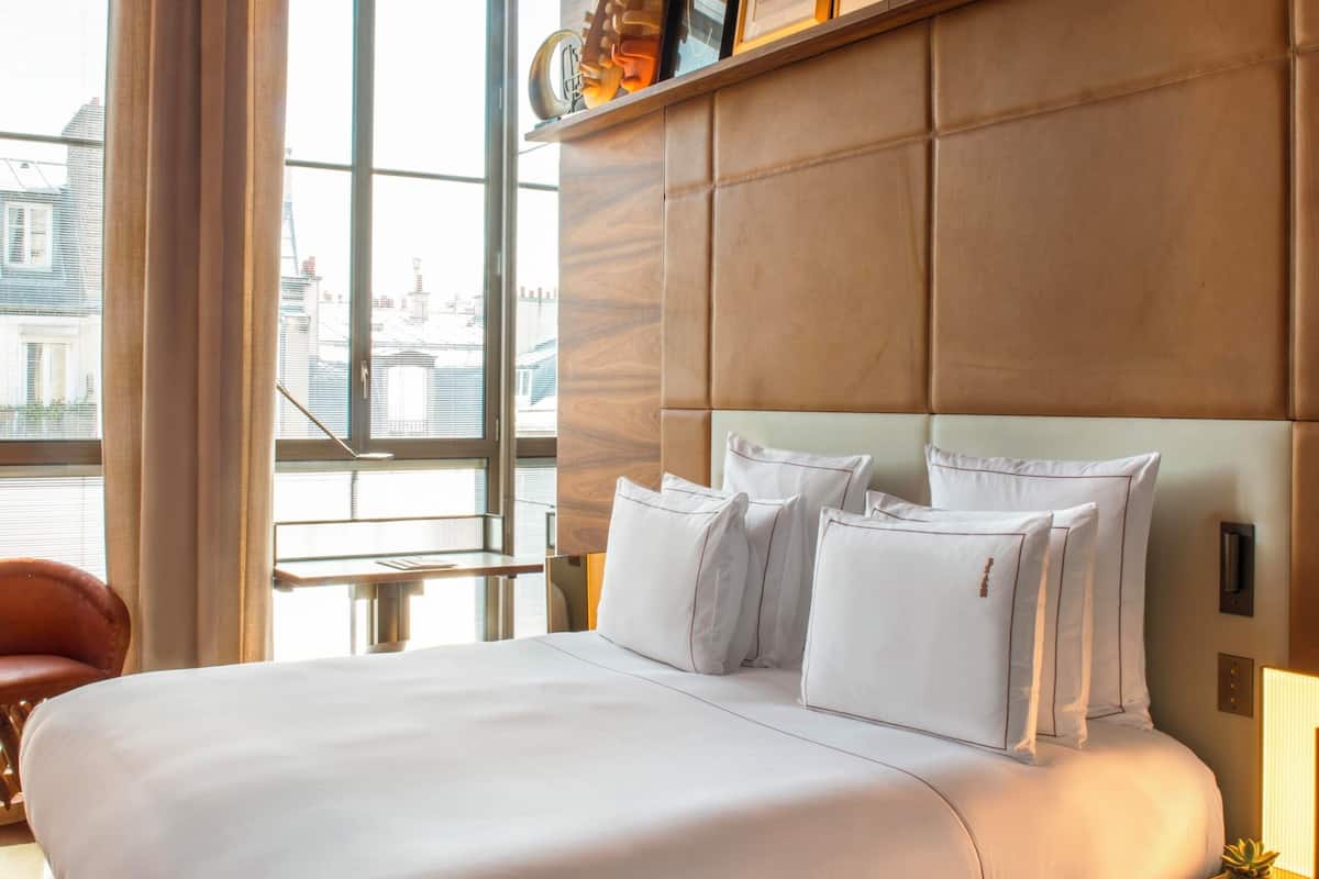 Habitación de hotel con una cama grande, cabecera con cuadros enmarcados, ventana con cortinas y vista a edificios.