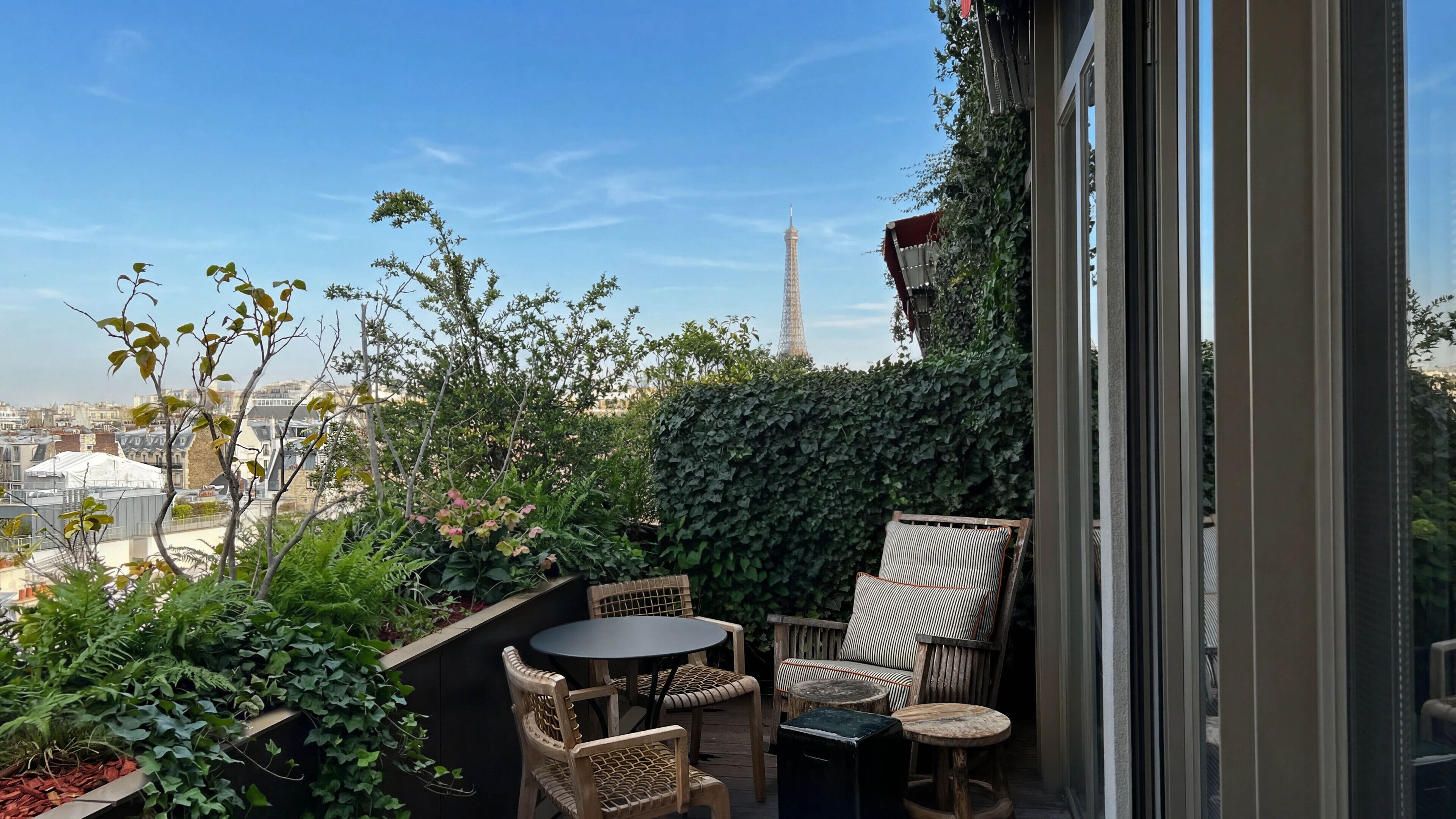A balcony with a view of the Eiffel Tower, surrounded by greenery and furnished with wicker chairs and a small table.
