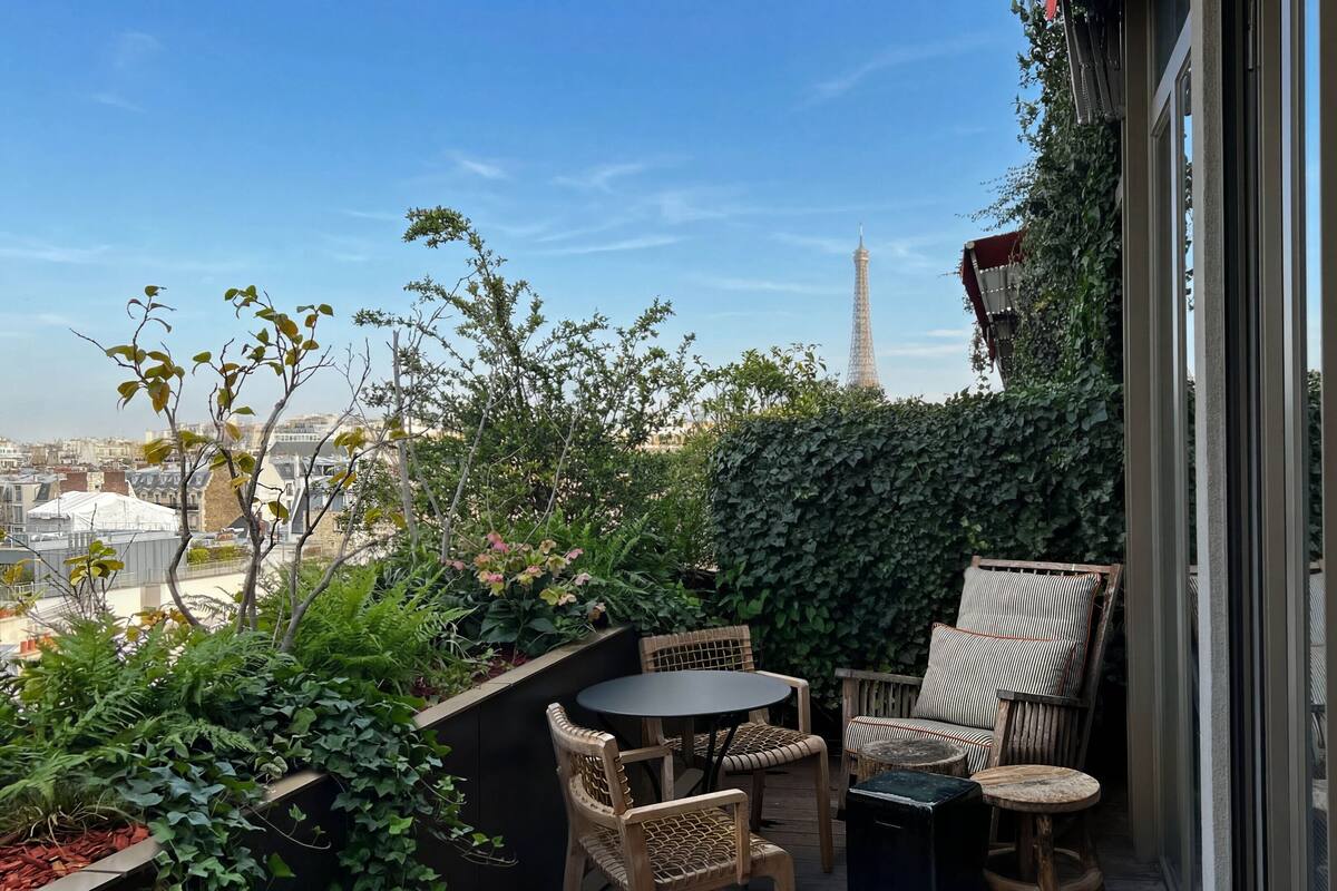 Un balcón con vistas a la Torre Eiffel, rodeado de vegetación y amueblado con sillas de mimbre y una pequeña mesa.