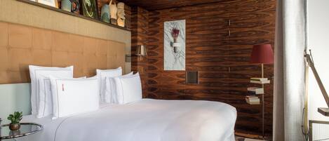 Una habitación de hotel moderna con una cama grande, paneles de madera, una mesita con una lámpara y una estantería.