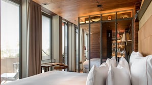 Ein modernes Hotelzimmer mit einem großen Bett, einem kleinen Tisch, einem Stuhl und Blick auf die Stadt durch große Fenster.