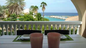 Un balcon avec une table dressée pour deux personnes, donnant sur une zone côtière bordée de palmiers et offrant une vue dégagée sur la mer.