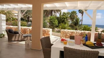 Uma área de estar coberta ao ar livre com móveis de vime, uma mesa posta com frutas e bebidas, e vista para o oceano e palmeiras.