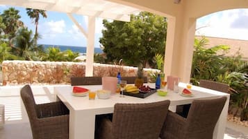 Uma área de refeições ao ar livre com cadeiras de vime e uma mesa branca posta com frutas, bebidas e uma melancia.