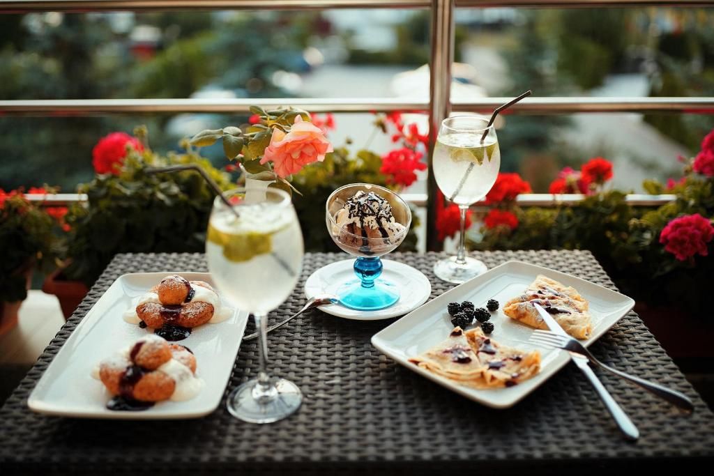 Ett bord med en desserttallrik, ett glas med en ljus dryck och en dessert i en blå skål, placerat framför ett fönster med utsikt över grönska och blommor.
