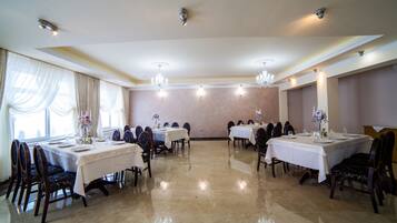 Uma sala de jantar formal com várias mesas postas para refeição, elegantes lustres e uma grande janela com cortinas.